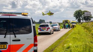 Op een smalle landweg staat een rij met politie-auto's met in de verte een ambulance waar hulpverleners bezig zijn rond een brancard en in de lucht komt een traumaheli aanvliegen