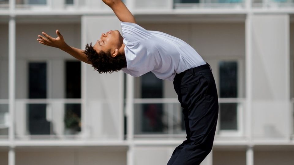 Danser in zwarte broek en wit shirt buigt achterover, op de achtergrond de balkonnetjes van een flatgebouw
