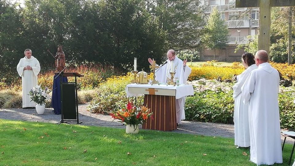 Eucharistieviering in de open lucht met pastoor achter altaar naast het gazon met volwassen misdienaars aan weerszijden
