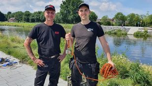 Twee mannen gekleed in zwarte broeken, zwarte t-shirts en basebalpetjes staan voor een brede sloot en één van hen houdt een oranje touw omhoog met een magneet er aan