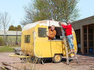 Gele caravan wordt geschuurd door vrijwilligers: eentje hangt uit het raam, eentje staat op een ladder tegen de buitenkant van de caravan
