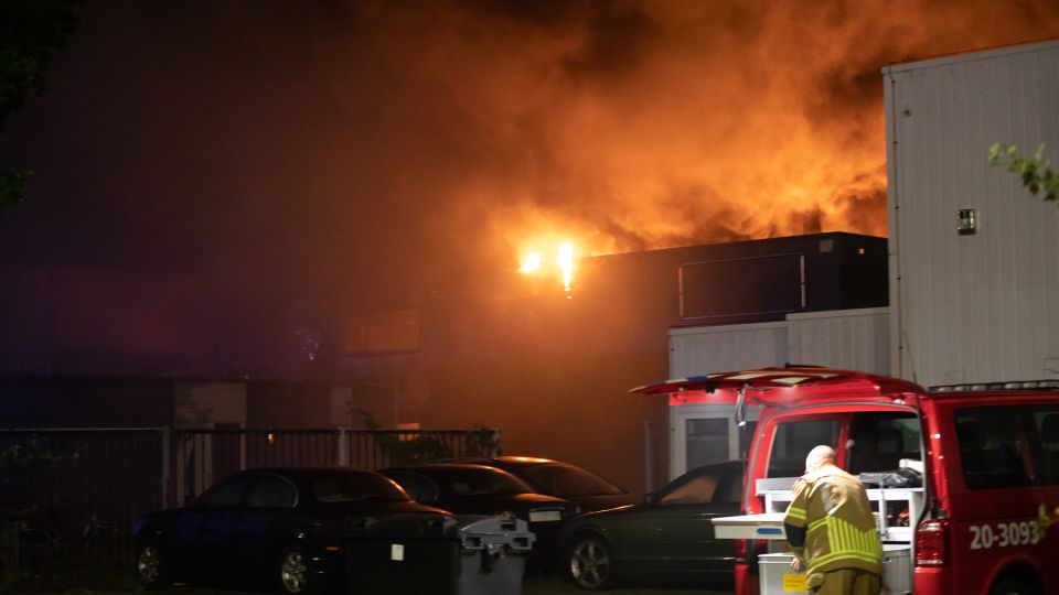 Brandweerman haalt spullen uit een brandweerbus terwijl op de achtergrond de vlammen uit het dak van een bedrijfspand slaan
