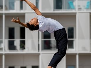 Danser in zwarte broek en wit shirt buigt achterover, op de achtergrond de balkonnetjes van een flatgebouw