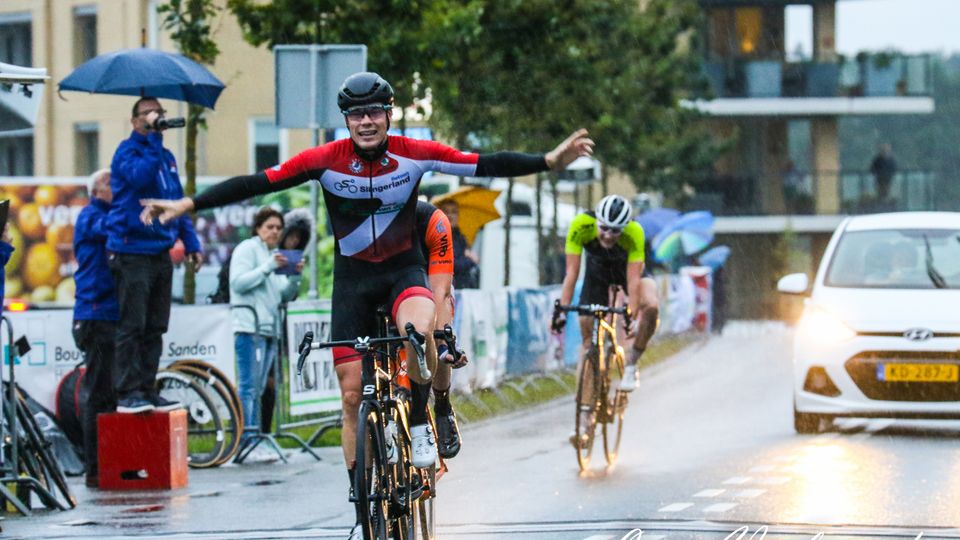 Wielrenner Remco Schouten rijdt in rood-wit-zwart shirt met armen wijd over finish in Hoogerheide langs publiek met paraplu's tegen de regen